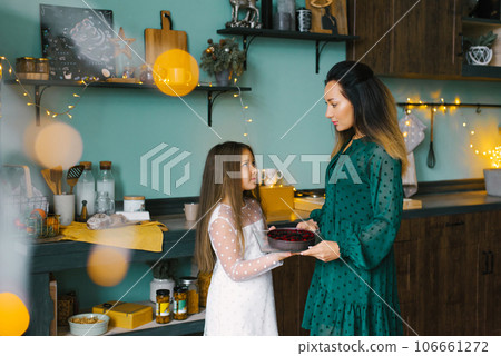 Mom is holding a pie with berries in her hands, daughter is enjoying the Christmas holiday at home Mom is holding a pie with berries in her hands, daughter is enjoying the Christmas holiday at home 106661272