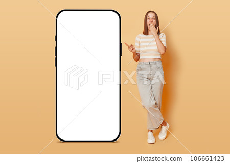 Surprised Caucasian woman wearing casual attire standing near big huge blank screen of mobile phone with advertisement area point finger at blank display isolated over beige background. 106661423