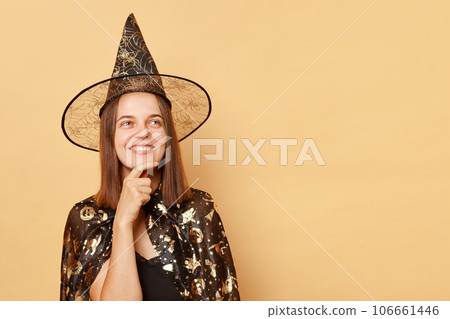 Dreamy young girl wearing black halloween garment and cone posing isolated over beige background looking smiling away at copy space for advertisement Dreamy young girl wearing black halloween garment and cone posing isolated over beige background looking smiling away at copy space for advertisement 106661446