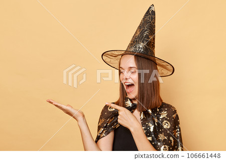 Amazed cheerful young girl wearing black halloween garment and cone posing isolated over beige background presenting copy space on palm pointing at advertisement area. Amazed cheerful young girl wearing black halloween garment and cone posing isolated over beige background presenting copy space on palm pointing at advertisement area. 106661448