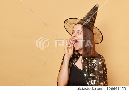 Cheerful young girl wearing black halloween garment and cone posing isolated over beige background keeps hand near mouth screaming making announcement 106661453