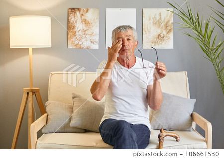 Tired senior with glasses. Sore eyes from infection. mature old man wearing white T-shirt rubbing and wiping eyes sitting on sofa in living room home interior Tired senior with glasses. Sore eyes from infection. mature old man wearing white T-shirt rubbing and wiping eyes sitting on sofa in living room home interior 106661530