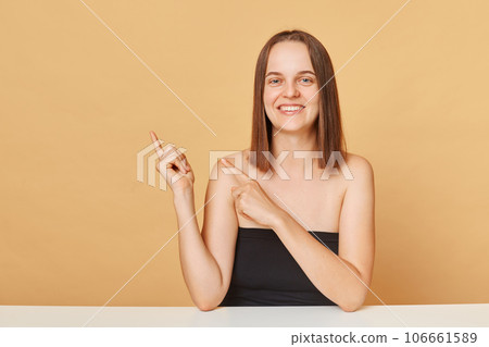 Happy young happy woman wearing black top looking at camera posing isolated over beige background pointing at space for promotional text advertisement area mockup Happy young happy woman wearing black top looking at camera posing isolated over beige background pointing at space for promotional text advertisement area mockup 106661589