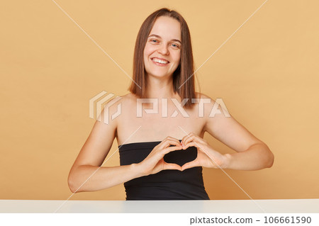Cute smiling cheerful young happy woman wearing black top looking at camera posing isolated over beige background showing heart shape with hands expressing love. Cute smiling cheerful young happy woman wearing black top looking at camera posing isolated over beige background showing heart shape with hands expressing love. 106661590
