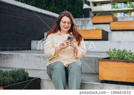 Smiling woman text messaging on smart phone at sunny day. Urban lifestyle concept. Traveler sitting and reading news on smartphone, fast connection, checking mobile apps outdoors. 106661610