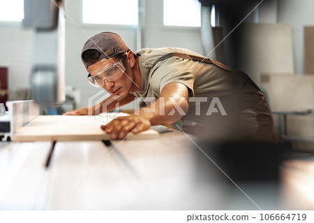 Young carpenter cutting a piece of wood in using a circular saw in furniture factory 106664719