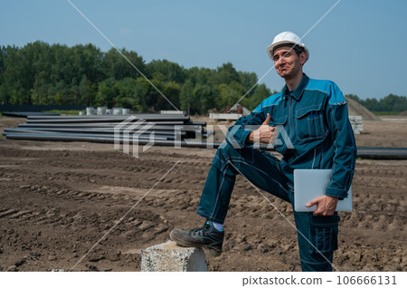Construction worker holding laptop and showing thumb up while standing at construction site.  106666131