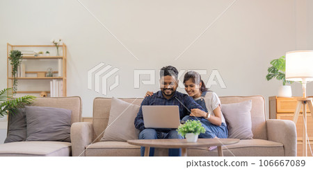 Happy indian family couple using laptop looking at computer laptop sitting on sofa together relaxing at home Happy indian family couple using laptop looking at computer laptop sitting on sofa together relaxing at home 106667089
