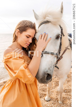A white horse and a woman in a dress stand on a beach, with the sky and sea creating a picturesque backdrop for the scene. A white horse and a woman in a dress stand on a beach, with the sky and sea creating a picturesque backdrop for the scene. 106667521