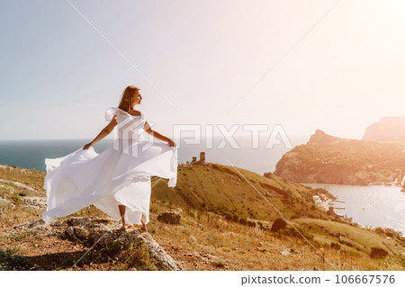 Happy woman in a white dress and hat stands on a rocky cliff above the sea, with the beautiful silhouette of hills in thick fog in the background. Happy woman in a white dress and hat stands on a rocky cliff above the sea, with the beautiful silhouette of hills in thick fog in the background. 106667576