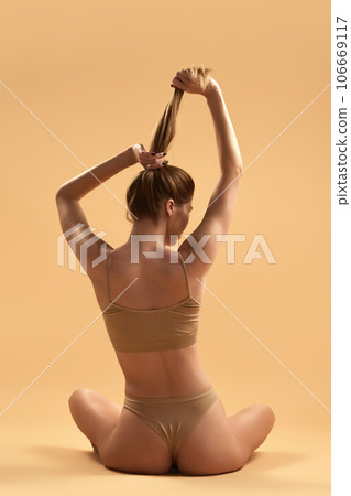 Rear view of young girl with slim body, buttocks sitting in lotus pose in beige underwear against sand studio background. healthy back. Rear view of young girl with slim body, buttocks sitting in lotus pose in beige underwear against sand studio background. healthy back. 106669117