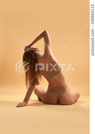Rear view of young girl with slim body sitting in lotus pose in beige inner wear against sand studio background 106669118