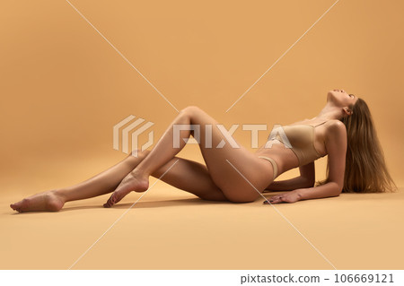 Full lenght portrait of young thin girl with short hair wearing beige underwear lying on floor over sand color background. Full lenght portrait of young thin girl with short hair wearing beige underwear lying on floor over sand color background. 106669121