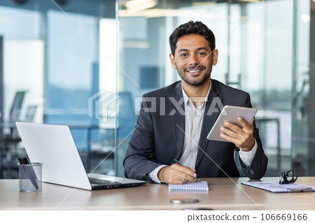 Portrait of young businessman with tablet computer inside office, hispanic man smiling and looking at camera, working with laptop, using app on tablet. Portrait of young businessman with tablet computer inside office, hispanic man smiling and looking at camera, working with laptop, using app on tablet. 106669166