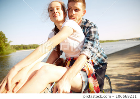 Portrait of young cheerful, lovely couple enjoying, having fun cycle ride over river embankment in hot summer day in evening. Portrait of young cheerful, lovely couple enjoying, having fun cycle ride over river embankment in hot summer day in evening. 106669211
