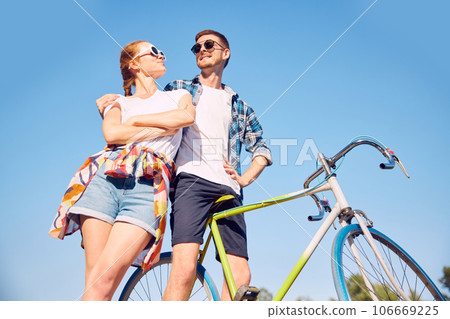 Bottom view photo of stylishly dressed couple with bicycle in summer sunlight in park. Two friends looking with love each other walking near river with cycles. 106669225