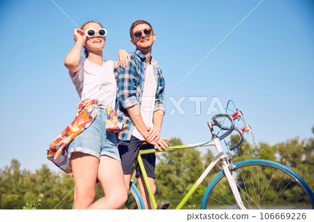 Bottom view photo of stylishly dressed couple walking with bicycle in summer sunlight park feel love. Two friends looking with love each other walking with cycles. 106669226