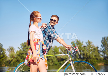 Side view photo of couple walking with bicycle in summer sunlight garden feel cherish love. Two friends looking with love each other walking near river with cycles. 106669227