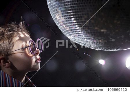A young boy with glasses looks at a spinning disco ball. Children's disco, party. A young boy with glasses looks at a spinning disco ball. Children's disco, party. 106669510