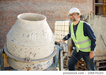 Worker standing wearing white helmet. Man standing near concrete mixer. Construction site on a background. 106670283