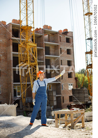 Construction site manager standing wearing orange helmet. Man giving an order to workers. Construction site on a background. 106670302