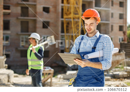 Architect watching some details on a construction. Worker standing at building site with construction structure in background. Man in background are blurred. Architect watching some details on a construction. Worker standing at building site with construction structure in background. Man in background are blurred. 106670312