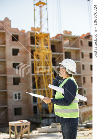 Construction site manager standing wearing safety vest and helmet, thinking at construction site. Architect with papers at a construction site. Man talking on the phone. 106670324