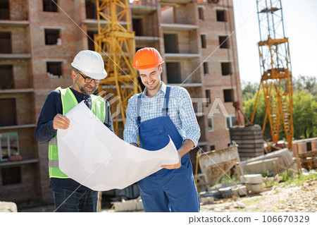 Worker and architect watching some details on a construction. Two engineers talking at building site with construction structure in background. Men wearing helmets. Worker and architect watching some details on a construction. Two engineers talking at building site with construction structure in background. Men wearing helmets. 106670329