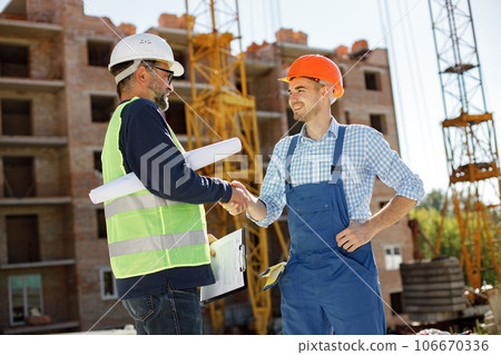 Worker and architect watching some details on a construction. Two engineers talking at building site with construction structure in background. Men wearing helmets. Worker and architect watching some details on a construction. Two engineers talking at building site with construction structure in background. Men wearing helmets. 106670336