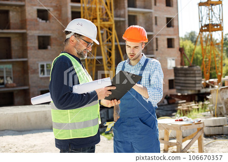 Worker and architect watching some details on a construction. Two engineers talking at building site with construction structure in background. Men wearing helmets. Worker and architect watching some details on a construction. Two engineers talking at building site with construction structure in background. Men wearing helmets. 106670357