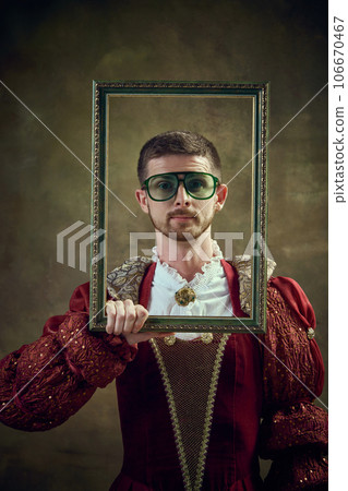 Portrait of medieval royal person, prince in female dress holding picture frame and looking against dark green background 106670467