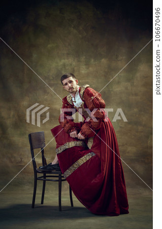 Young medieval prince, royal person leaning on chair, wearing female dress against dark green background. Renaissance style 106670496