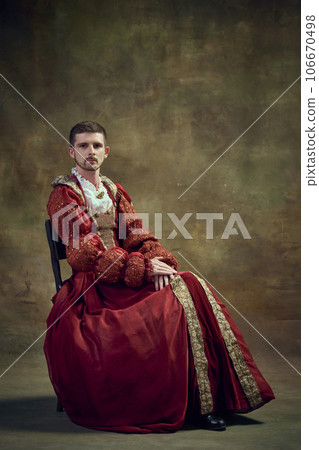Historical portrayal. Elegant young medieval prince sitting tenderly in female dress against dark green background Historical portrayal. Elegant young medieval prince sitting tenderly in female dress against dark green background 106670498