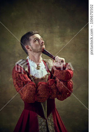 Thoughtful young man, medieval prince in female dress with fan against vintage green background. Equality 106670502