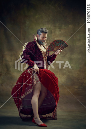 Drama. Humorous representation of medieval royal person, man in female dress with fan against vintage green background 106670518