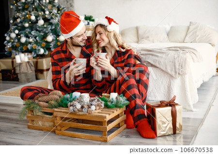 Romantic couple celebrating Christmas on a floor near Christmas tree. Blonde woman and brunette man wearing plaid pajamas. Romantic couple drinking a tea from a mugs. 106670538