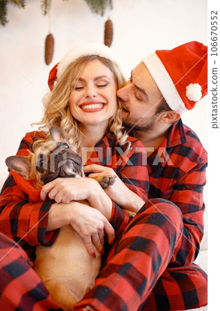 Portrait of lovely couple celebrating christmas with dog. Blonde woman and brunette man wearing plaid pajamas. French bulldog with his family. Portrait of lovely couple celebrating christmas with dog. Blonde woman and brunette man wearing plaid pajamas. French bulldog with his family. 106670552