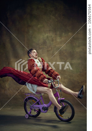 Young man, medieval royal person in female dress riding little bike against vintage background. Humorous emotions. Speed Young man, medieval royal person in female dress riding little bike against vintage background. Humorous emotions. Speed 106670564