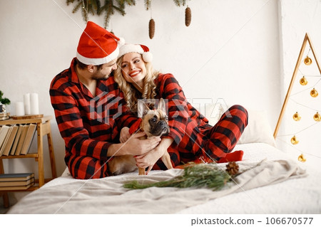 Lovely couple celebrating christmas with dog on a bed. Blonde woman and brunette man wearing plaid pajamas. French bulldog with his family. Lovely couple celebrating christmas with dog on a bed. Blonde woman and brunette man wearing plaid pajamas. French bulldog with his family. 106670577