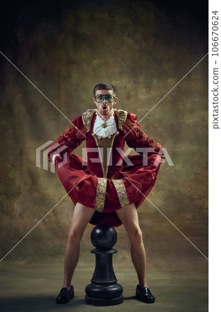 Emotional man, medieval royal person in female dress standing over giant chess piece against vintage green background. Game on Emotional man, medieval royal person in female dress standing over giant chess piece against vintage green background. Game on 106670624