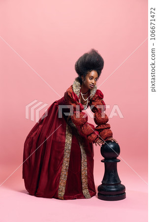 Checkmate. Portrait of medieval princess, queen in red costume playing with giant chess piece against pink studio background Checkmate. Portrait of medieval princess, queen in red costume playing with giant chess piece against pink studio background 106671472