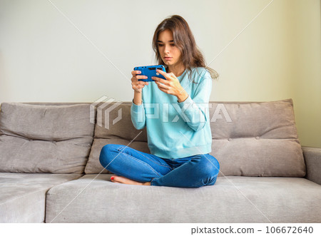 Surprised young woman holding smartphone with two hands,sitting on the sofa in casual clothes. She reading new or playing games. Lockdown activities Surprised young woman holding smartphone with two hands,sitting on the sofa in casual clothes. She reading new or playing games. Lockdown activities 106672640
