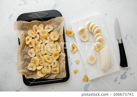Baking tray with dried banana chips and raw banana with kitchen knife.Top view. 106673741