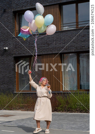 Woman in colored hair walks with an armful of balloons and drinks a refreshing beverage Woman in colored hair walks with an armful of balloons and drinks a refreshing beverage 106674525