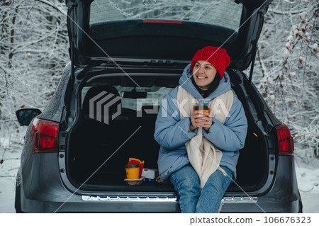 happy woman sitting in the trunk of her car in the winter forest and holding a coffee in her hand happy woman sitting in the trunk of her car in the winter forest and holding a coffee in her hand 106676323