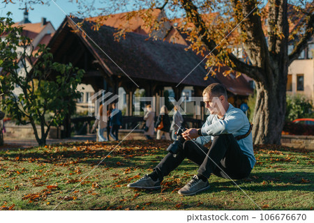 Professional Photographer Taking Picture Of Beautiful Autumn Park. Man Professional Photographer Sit With Camera And With Smartphone In Autumn Park. Retouched, Vibrant Colors, Brownish Tones. 106676670