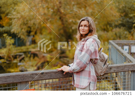 Portrait Of Cute Young Woman In Casual Wear In Autumn, Standing On Bridge Against Background Of An Autumn Park And River. Pretty Female Walking In Park In Golden Fall. Copy Space. Smiling Girl In The 106676721