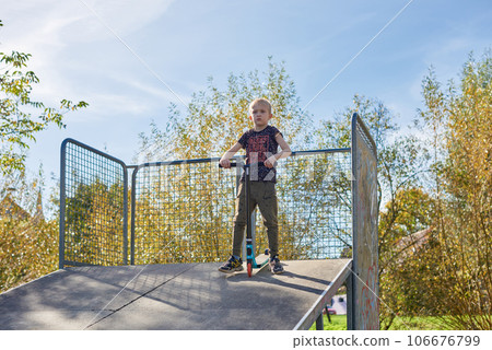 Boy on scooter makes a trick and enjoying his riding in the skate park at cloudy spring day. Young man doing trick on the kick scooter in the park. Full body little boy on the kick scooter on sunny Boy on scooter makes a trick and enjoying his riding in the skate park at cloudy spring day. Young man doing trick on the kick scooter in the park. Full body little boy on the kick scooter on sunny 106676799