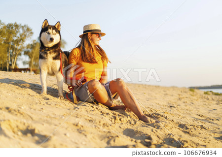 Woman in hat and sunglasses sits with her dog on the beach and enjoying sunset Woman in hat and sunglasses sits with her dog on the beach and enjoying sunset 106676934