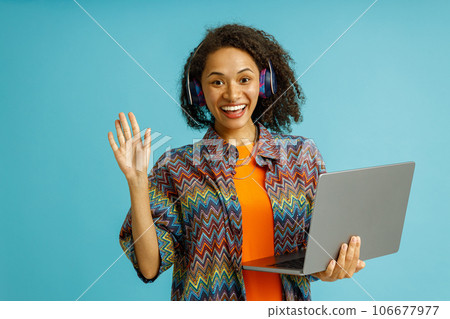 Smiling woman in headphones holding laptop and waving hi in camera on blue studio background 106677977